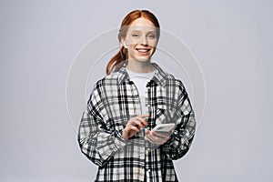 Attractive smiling redhead young woman wearing wireless headphones listening to music from smartphone.