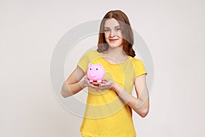 Attractive smiling teenager girl with brown hair holding piggy bank, being happy with money savings