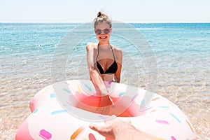 Attractive smiling woman is calling her boyfriend into the sea. Couple is holding doughnut inflatable ring. Follow me concept