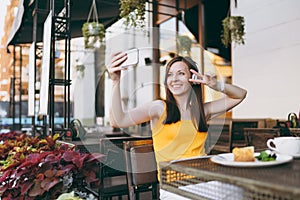 Attractive smiling woman in outdoors street coffee shop cafe sitting at table, listen music in headphones, doing selfie