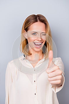 Attractive smiling woman in shirt giving a wink and showing thumb up on gray background