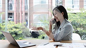 Attractive smiling young asian business woman relaxing at office, working on laptop computer, talking on mobile phone