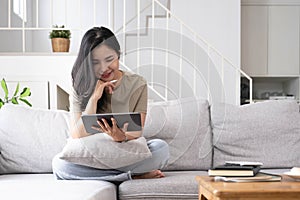 Attractive smiling young asian woman relaxing on a leather couch at home, working on laptop computer.