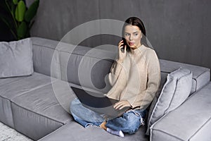 Attractive young business woman relaxing on a leather couch at home, working on laptop computer, talking on mobile phone