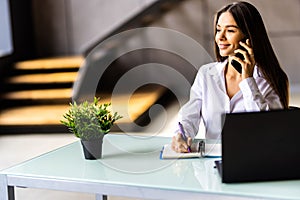 Attractive smiling young businesswoman wearing jacket talking on mobile phone while sitting on a desk and using laptop in office