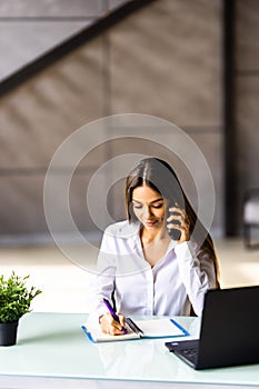 Attractive smiling young businesswoman wearing jacket talking on mobile phone while sitting on a desk and using laptop in office
