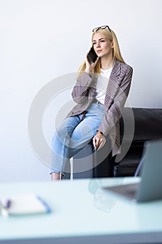 Attractive smiling young businesswoman wearing jacket talking on mobile phone while using laptop computer in office