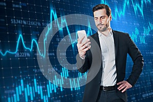 Attractive smiling young european businessman with cellphone and creative glowing forex chart on blurry background. Trade and