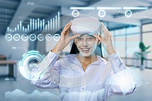 Attractive smiling young european businesswoman with VR glasses and glowing business chart hologram on blurry office interior