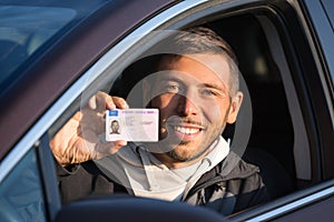 Attractive smiling young man proudly showing his driving license out of car window. Man has got driving license and feels very hap