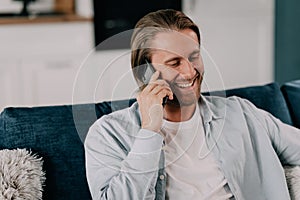 attractive smiling young man talking on mobile phone wearing casual clothes sitting on a couch at the living room