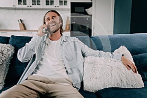 attractive smiling young man talking on mobile phone wearing casual clothes sitting on a couch at the living room