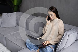 Attractive smiling young woman relaxing on a leather couch at home, working on laptop computer, talking on mobile phone
