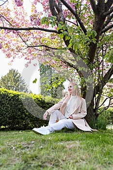 Attractive smiling young woman sitting in blossoming park on the lawn , warm spring day. Attractive girl on rest. Vertical frame