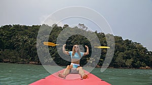 Attractive sportive blonde woman with sunglasses rows pink plastic kayak putting up paddle on sea against hills with