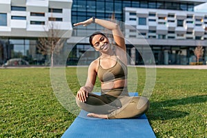 Attractive sporty black woman sitting on yoga mat, doing stretching exercses at urban park, empty space