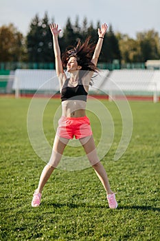 Attractive sporty brunette woman in pink shorts and top doing workout with jump rope in sun rays at the stadium