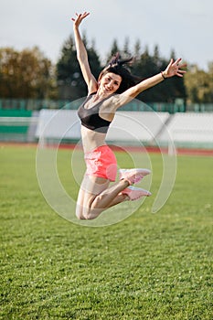 Attractive sporty happy brunette woman in pink shorts and top makes a high jump in sun rays at the stadium looking at the camera