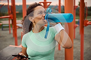 Attractive sporty Latin American woman with athletic physique holding a smartphone in one hand and drinking drinking water from a