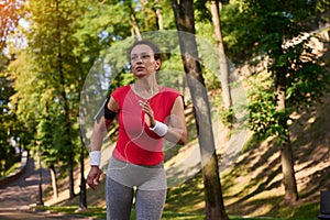 Attractive strength muscular multi ethnic woman in tight sportswear with earphones and smartphone holder running on the footpath