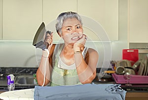 Attractive and stressed Asian middle aged lady ironing at home kitchen desperate and overwhelmed feeling unhappy in helpless face