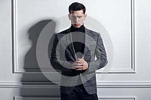 Frontal portrait of a handsome, elegant young man posing confident in stylish black-gray suit near white wall, studio.