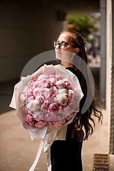 Attractive stylish brunette girl with eyes closed posing with bouquet of pink delicete peonies in european street