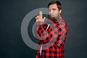 Attractive stylish hipster bearded macho man in red checkered shirt, making finger pistol gesture, standing as holding gun like