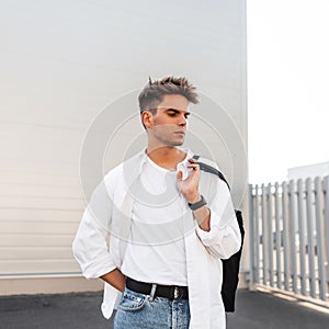 Attractive stylish young man in an elegant white shirt in blue jeans with a fashionable bag with a trendy hairstyle posing near a