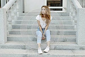 Attractive stylish young woman in a fashionable T-shirt in blue jeans in white trendy sneakers resting sitting on the steps