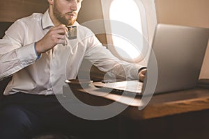 Attractive and successful businessman drinking coffee and working behind a laptop while sitting in a chair of his