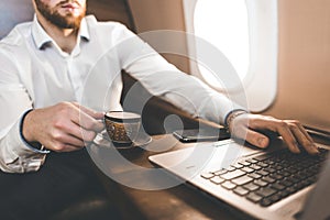 Attractive and successful businessman drinking coffee and working behind a laptop while sitting in a chair of his