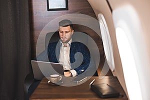 Attractive and successful businessman working on a laptop while sitting in a chair of his private jet.