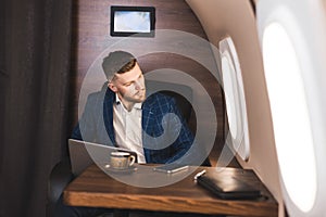 Attractive and successful businessman working on a laptop while sitting in a chair of his private jet.