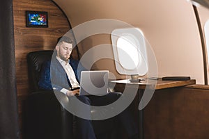 Attractive and successful businessman working on a laptop while sitting in a chair of his private jet.