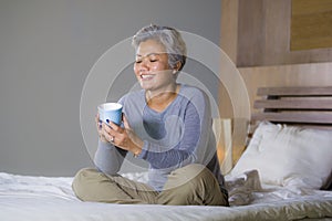 Attractive and successful mature woman with grey hair sitting on bed drinking coffee relaxed smiling happy as middle aged female