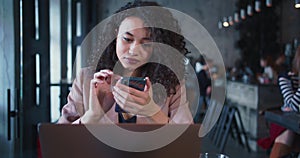 Attractive successful mixed race female IT manager with curly hair using smartphone app working online at trendy cafe.