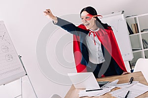 attractive super businesswoman in cape and mask screaming and standing with hand up