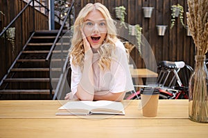 Attractive surprised blond girl amazedly looking in camera with book and coffee in courtyard of city cafe photo