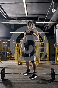 Attractive, tattooed, bearded man in black sport gloves, shorts, vest, cap and sneakers. Showing cool sign, barbell is