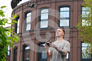 Attractive Tattooed Man Launching Drone Quadcopter and Looking At It. Stylish Red Bricked Building IS on Background