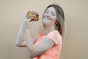 Attractive teen girl stands against the background of an orange wall in an orange T-shirt and holds a big burger in her hand, she