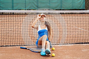 Attractive tennis player of European appearance in tennis clothes sitting on a tennis court. Posing near the tennis net. Resting
