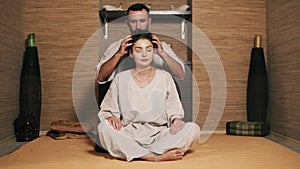 Attractive thai massagist is massaging young woman`s head sitting behind her. Beautiful girl with her eyes closed is