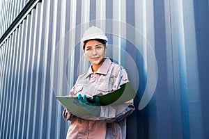 Attractive Thai woman industrial engineer in white hard hat, safety vest with a blurred container yard in background. Working.