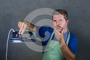 Attractive and thoughtful man holding clothes iron as if wondering a strange and weird device in messy husband or single guy doing