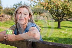 Attractive Thoughtful Middle Aged Woman Resting on Fence With Mug of Coffee or Tea