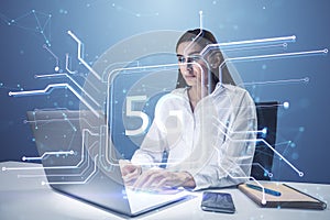 Attractive thoughtful young businesswoman using laptop at desk with creative square circuit button hologram with 5G icon.