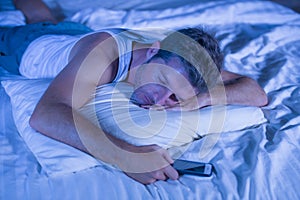 Attractive tired man in bed falling asleep while using mobile phone still holding the cellular in his hand while sleeping in inter
