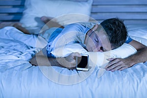 Attractive tired man in bed falling asleep while using mobile phone still holding the cellular in his hand while sleeping in inter